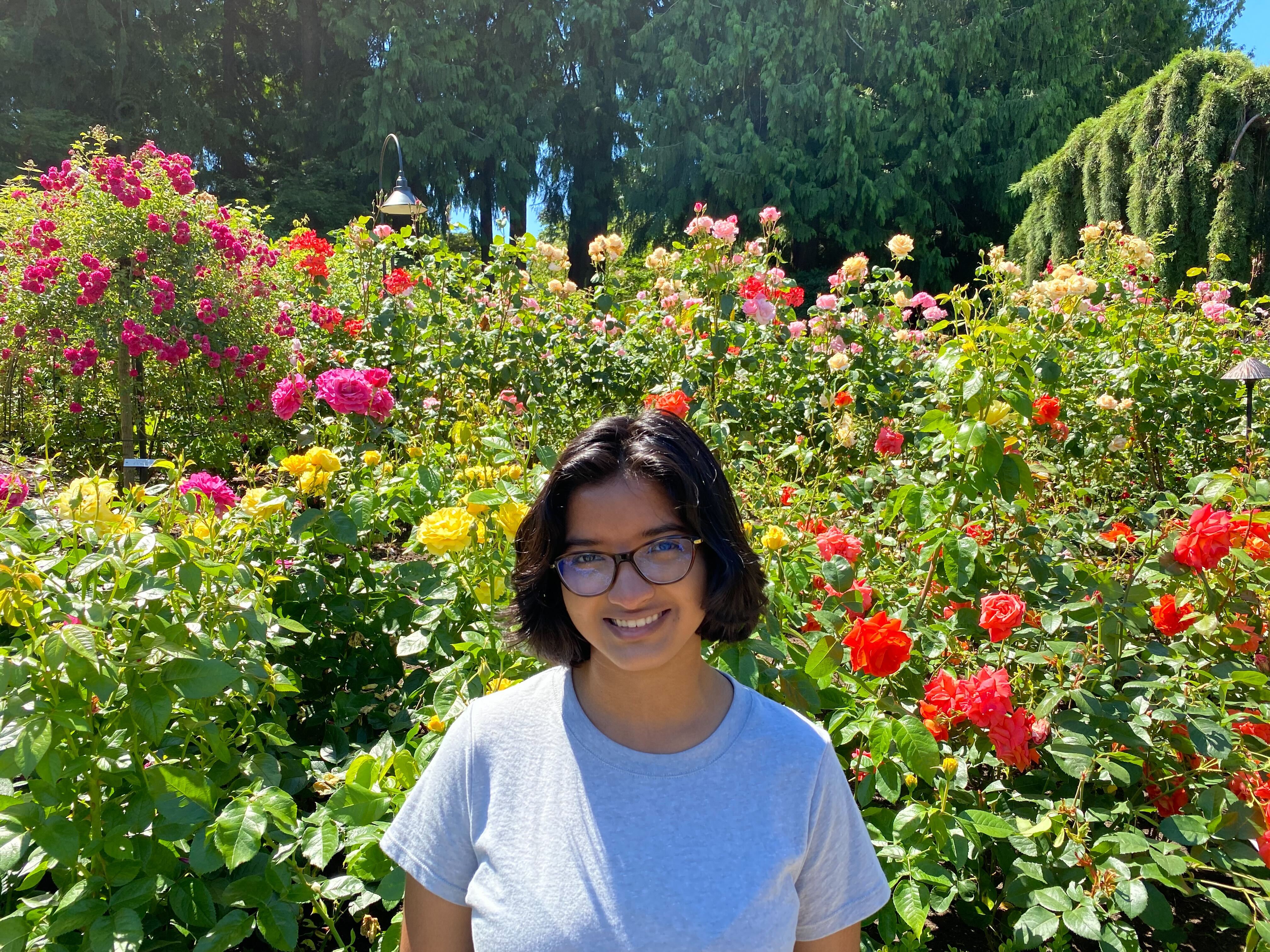 An Indian woman with short hair wearing a grey shirt is standing in front of flowers