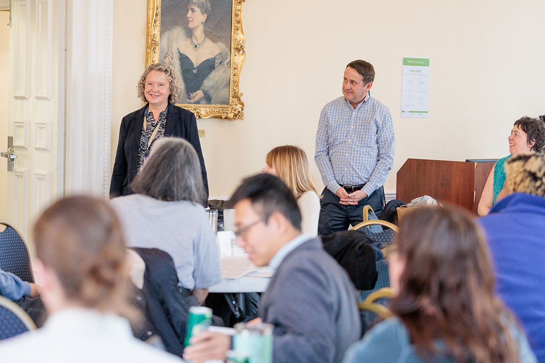 Two speakers stand at the front of a room with a group of writers listening at tables