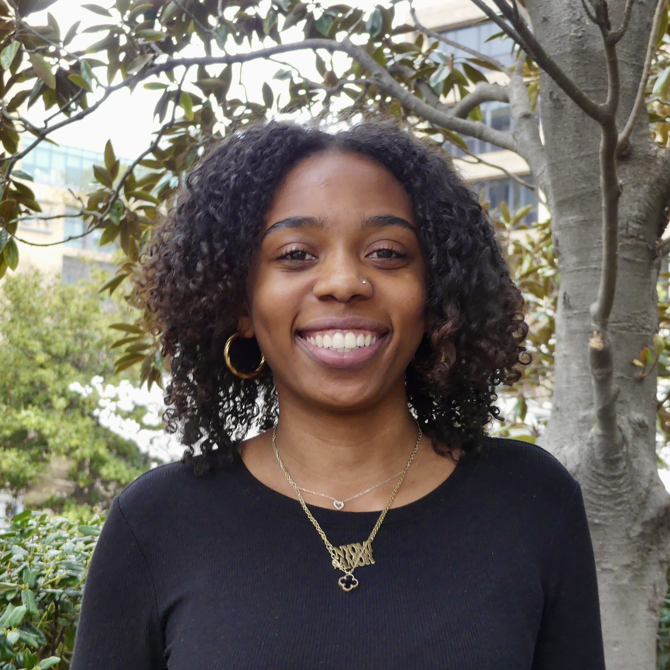 Nyomi, a young Black woman with curly hair and a black top, with brown eyes, smiling. 