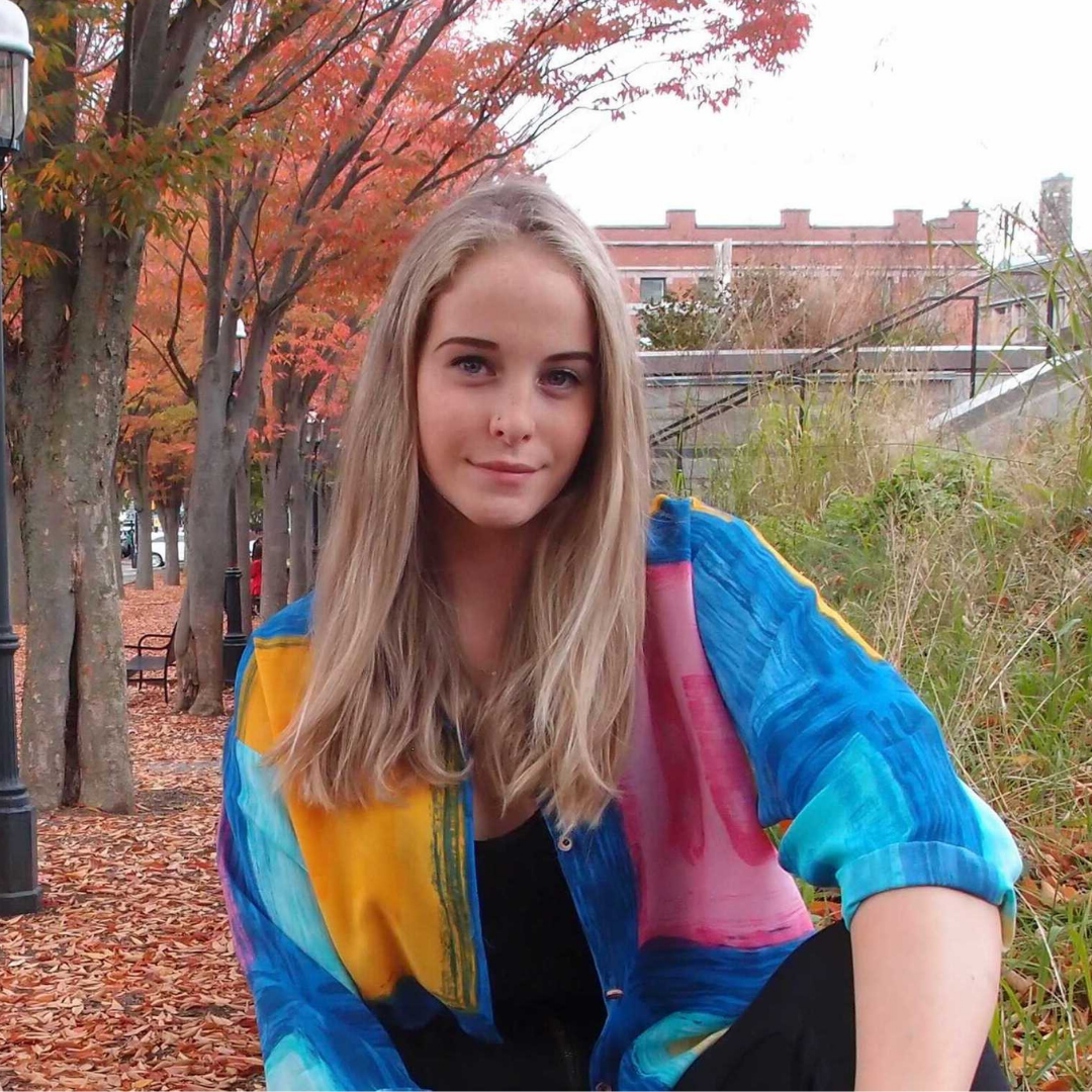 A young white woman is sitting, facing the camera and smiling. She has medium length blonde hair, blue eyes, and is wearing a blue, pink, and yellow blouse and a black undershirt. Behind her are red and orange autumn foliage and grass.
