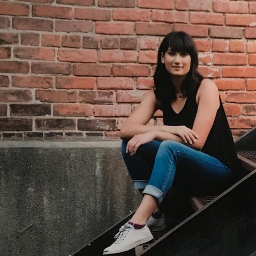 A young multiracial woman is sitting on an outdoor staircase looking at the camera with a slight smile. She has shoulder length, dark brown hair with bangs. She is wearing jeans, a black tank top, and grey converse shoes.