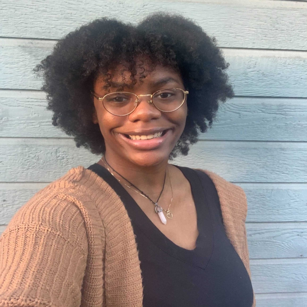 Kam, a young African American woman with glasses is smiling at the camera. She has a medium length curly hair, and is wearing a brown sweater with a black shirt underneath. A blue wall is behind her.
