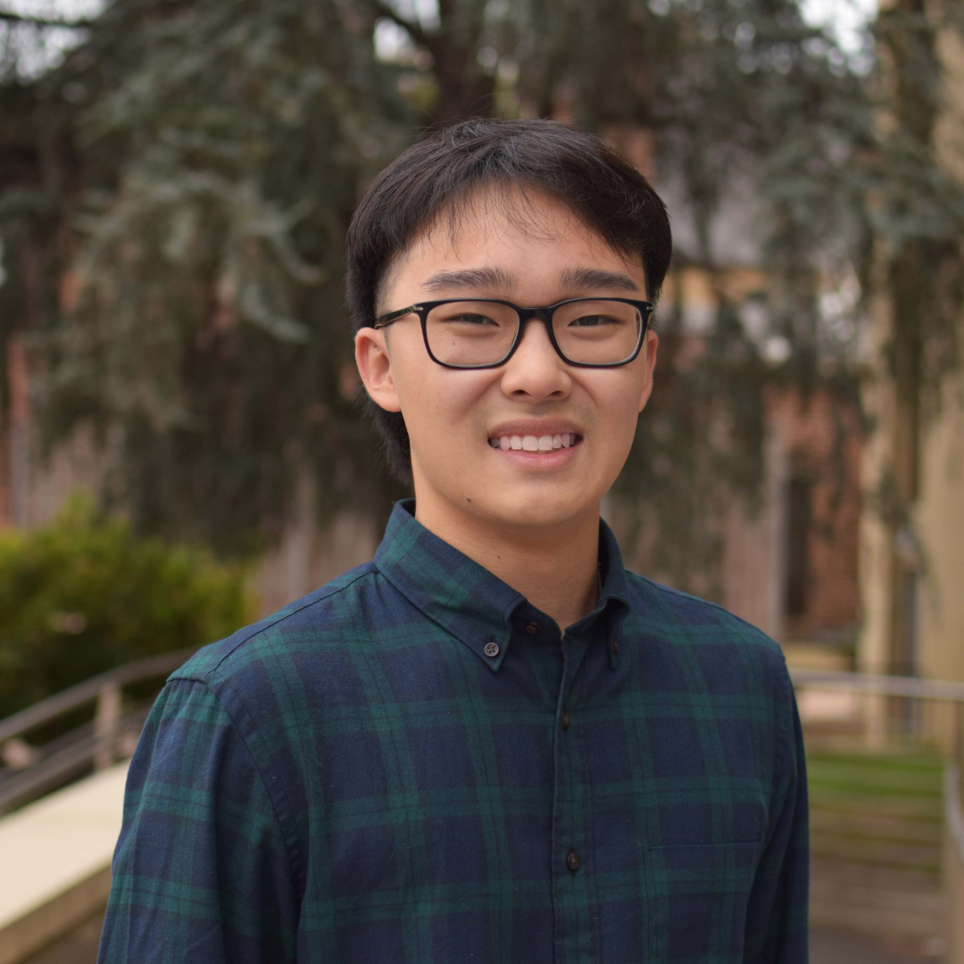 A young, Asian man smiling at the camera. He has black hair and black glasses. He's wearing a green and navy flannel. Behind him is blurred greenery.