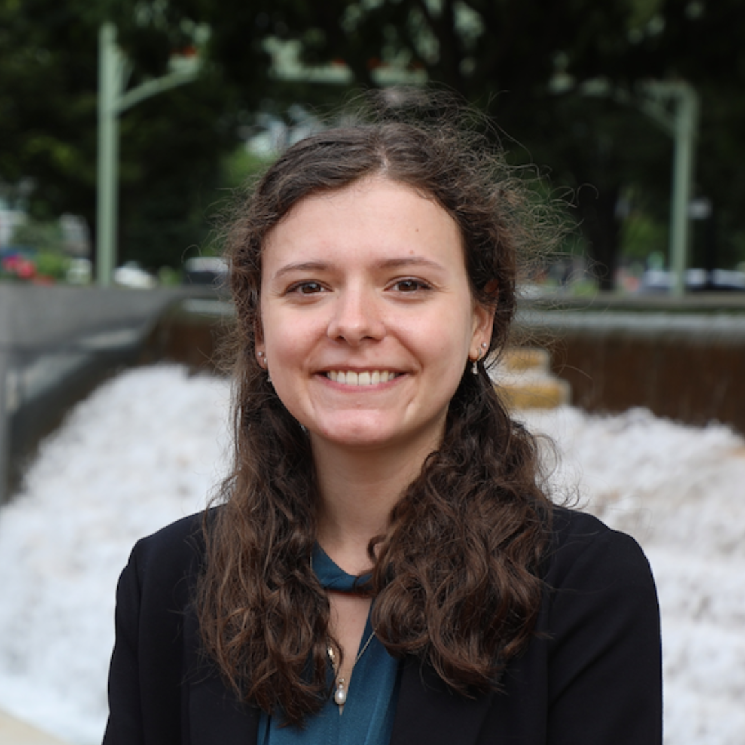 Morgan, a young white woman is facing the camera and smiling. She has medium length, curly brown hair and brown eyes. She is wearing a blue shirt with a black blazer and standing in front of a fountain and trees.