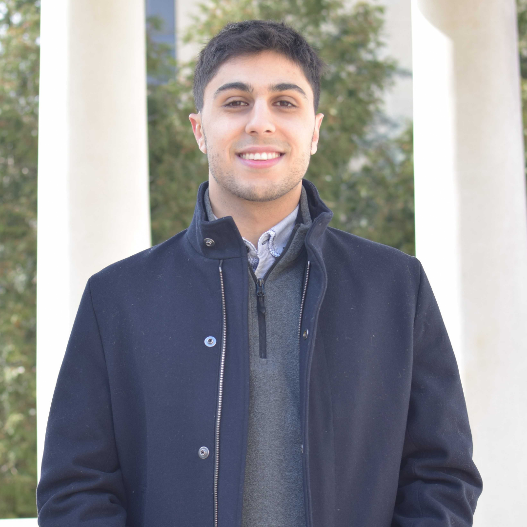 A young Persian man is smiling, looking directly at the camera. He has short, dark brown hair. He is wearing a dark navy jacket and standing in front of a pair of white Corinthian columns.
