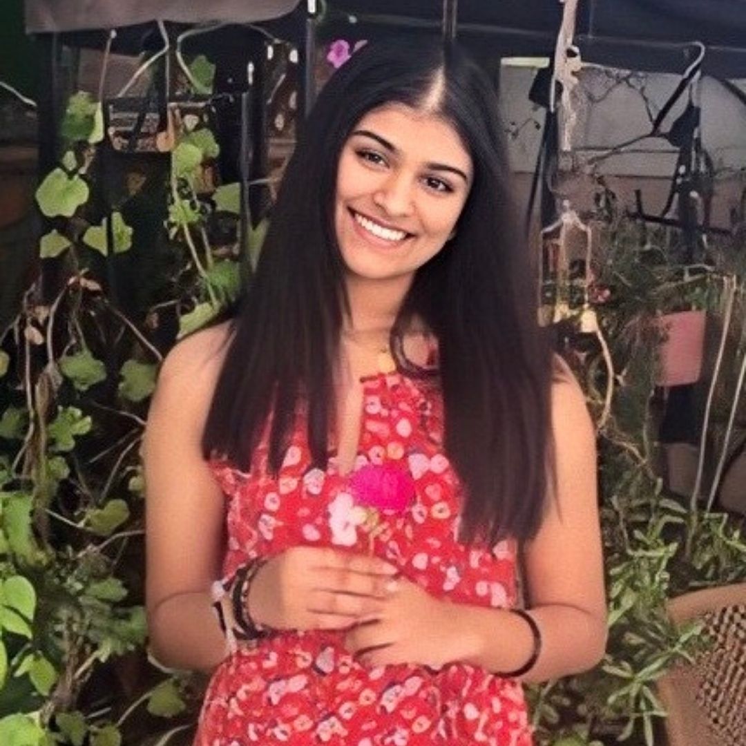 A young South Asian woman is smiling towards the camera with her head titled to the side. She is wearing a brightly colored floral dress and is standing in front of various plants and flowers.
