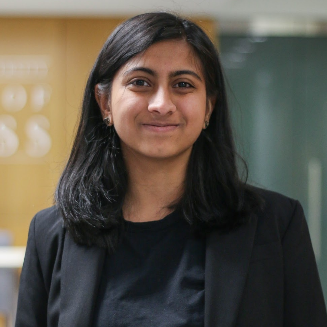 A young South Asian woman with medium-length straight black hair and brown eyes is smiling directly into the camera. She is wearing professional clothing, including a black shirt, black blazer, and round black earrings. She is in front of a blurred background.