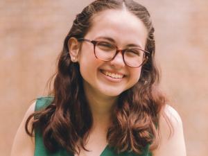 Lizzy, a young white women with wavy brown hair, brown eyes, and glasses, wearing a green jumpsuit, smiling