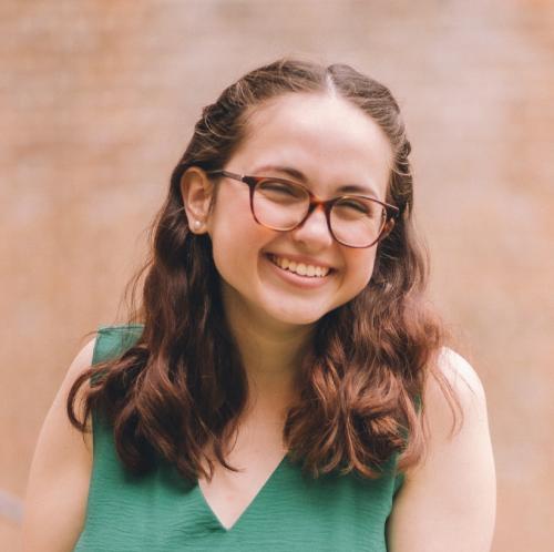 Lizzy, a young white women with wavy brown hair, brown eyes, and glasses, wearing a green jumpsuit, smiling