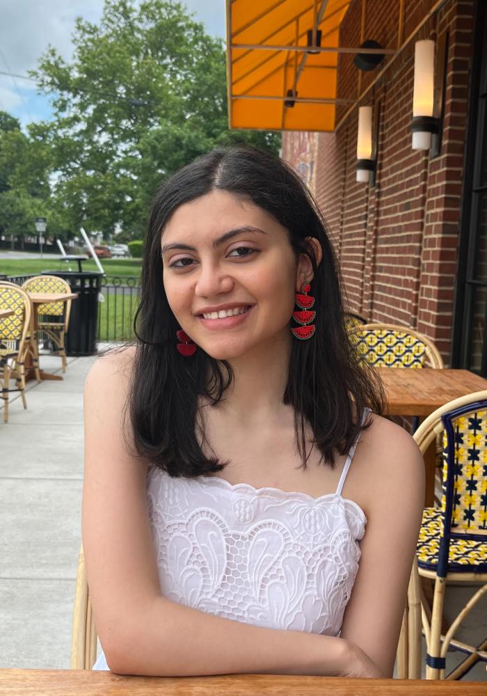 Nour, a young woman with dark brown hair and red earrings, smiles at the camera.
